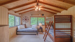 A cabin interior with a bunk bed, a single bed, a ceiling fan, a compass wall decoration, and a window with a view of trees.