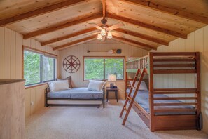 A cabin interior with a bunk bed, a single bed, a ceiling fan, a compass wall decoration, and a window with a view of trees.