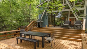 An outdoor deck with a table and chairs, surrounded by wooden railings and trees.
