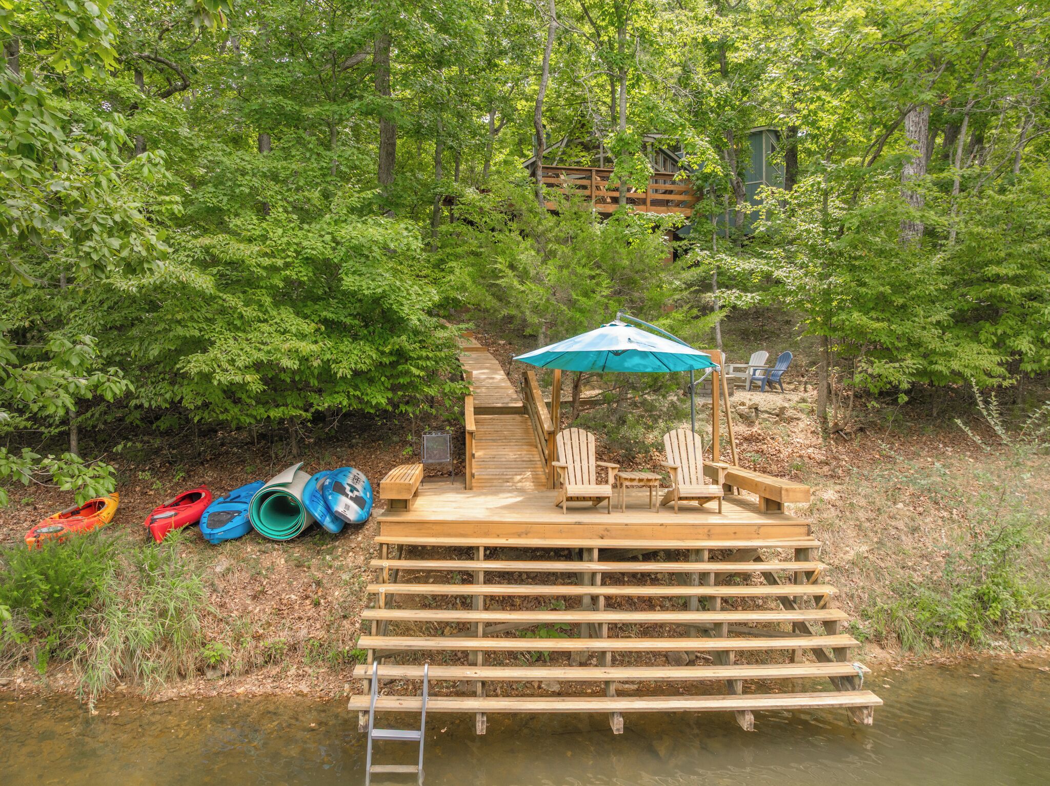 A wooden dock with a bench and a table, kayaks, and a paddleboard by a forested area.