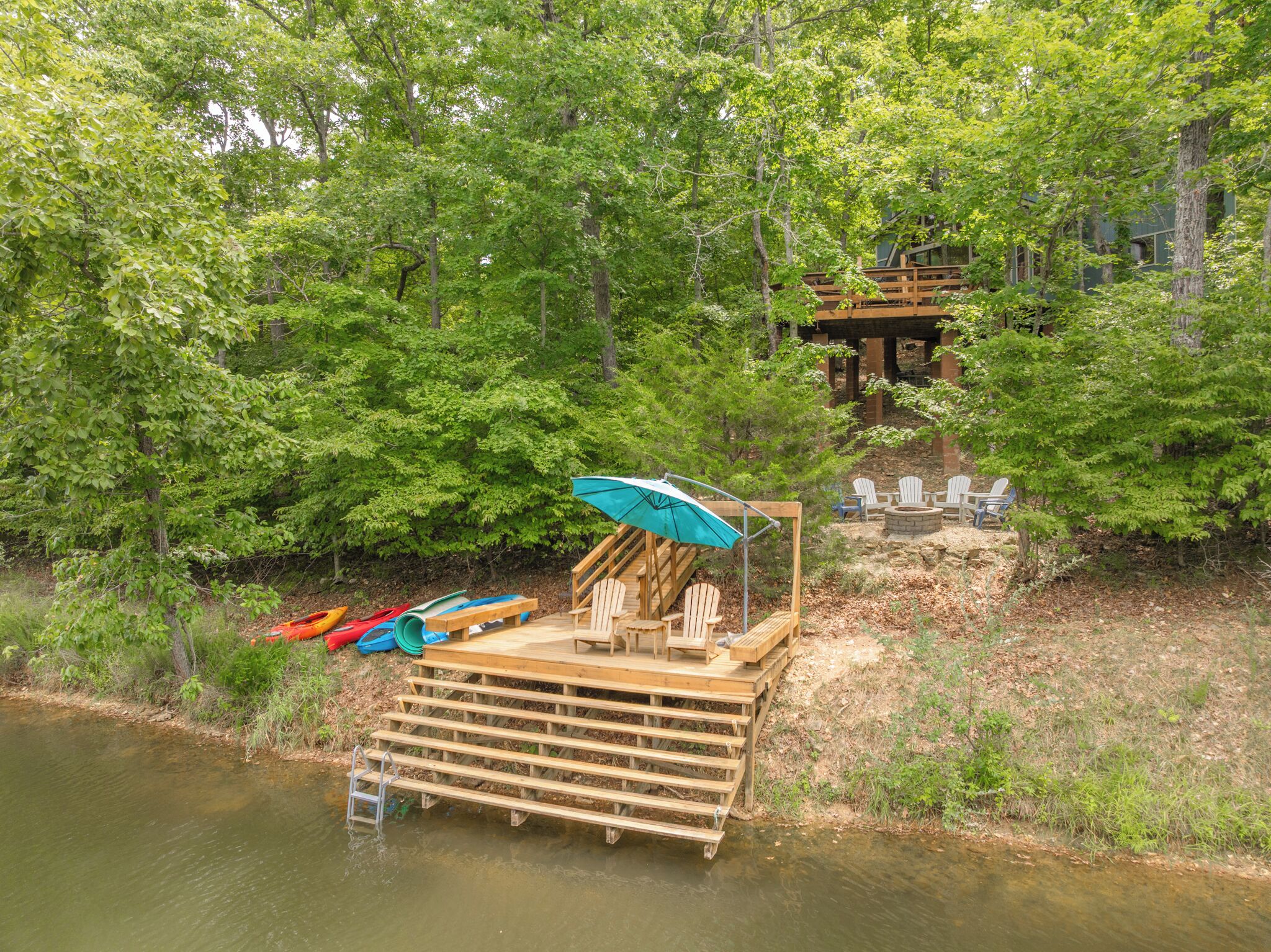 A wooden deck with chairs and a table, kayaks, and a small pavilion in a forested area by a river.
