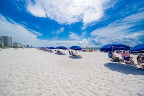 Une plage bordée de rangées de parasols bleus et de chaises longues, un ciel dégagé et des bâtiments en arrière-plan.