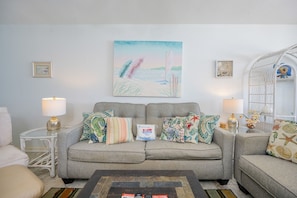 Ein Wohnzimmer mit grauem Sofa, bunten Kissen, einem Glastisch und einem Wandbild mit Strandmotiv.