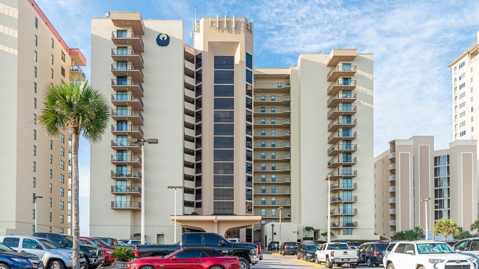 A multi-story hotel with a central tower, balconies, and a parking lot with various vehicles.