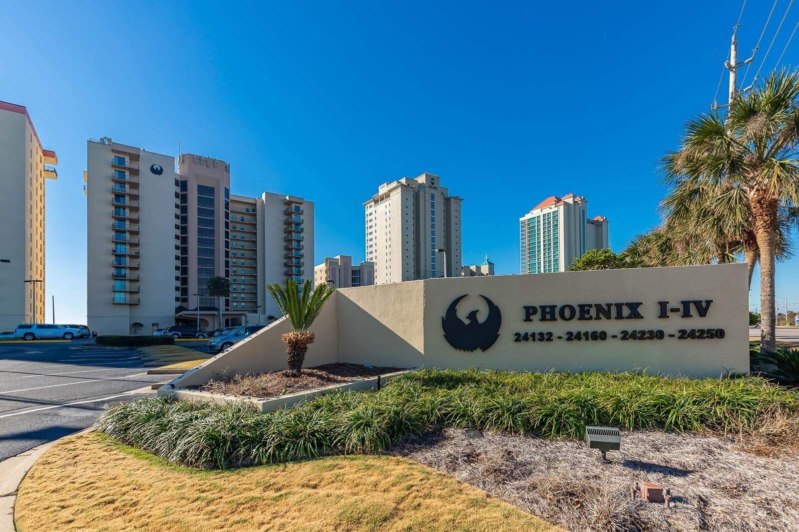 A sign for Phoenix I-IV with a bird logo, surrounded by a landscaped area with palm trees and a parking lot.