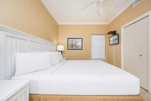Una habitación de hotel con cama, mesita de noche, lámpara, un cuadro, una ventana con persianas y una puerta.
