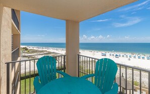 A balcony with two chairs and a table, overlooking a beach with umbrellas and people. - Phoenix V 414 1 Bedroom Condo (Orange Beach)