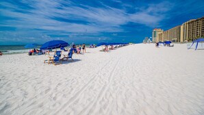 En strand med rader av blå parasoller och strandstolar, en sandstrand och en hotellbyggnad i bakgrunden.