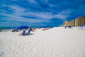 A beach with rows of blue umbrellas and beach chairs, a sandy shore, and a hotel building in the background. - Phoenix V 711 1 Bedroom Condo (Orange Beach)