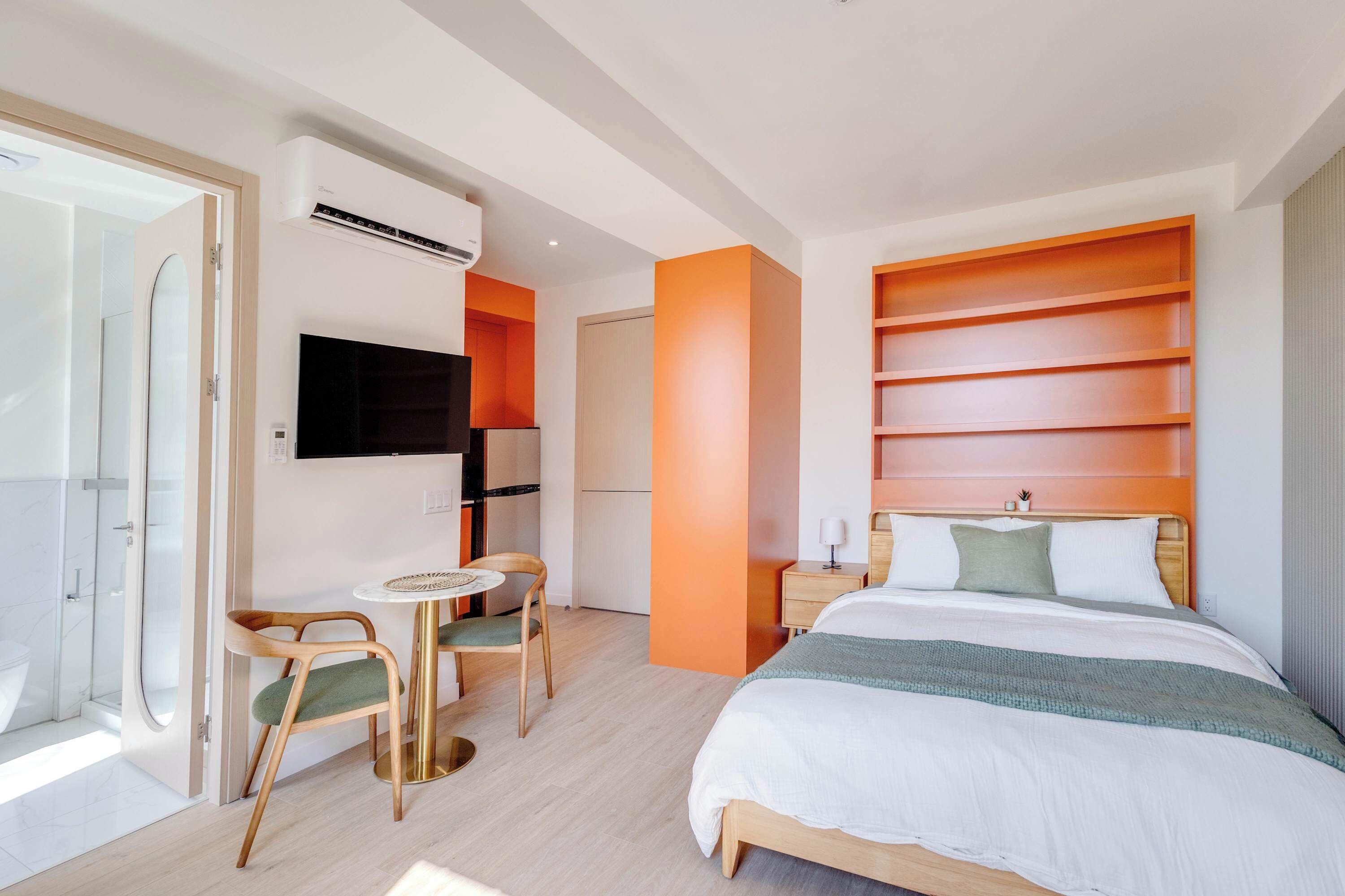 A modern hotel room with a bed, a TV, a small table with chairs, and a bathroom with a shower.