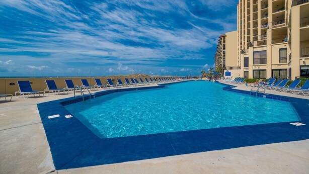 A large outdoor swimming pool with blue water, surrounded by lounge chairs and a tall building in the background. - Phoenix V 1111 1 Bedroom Condo (Orange Beach)