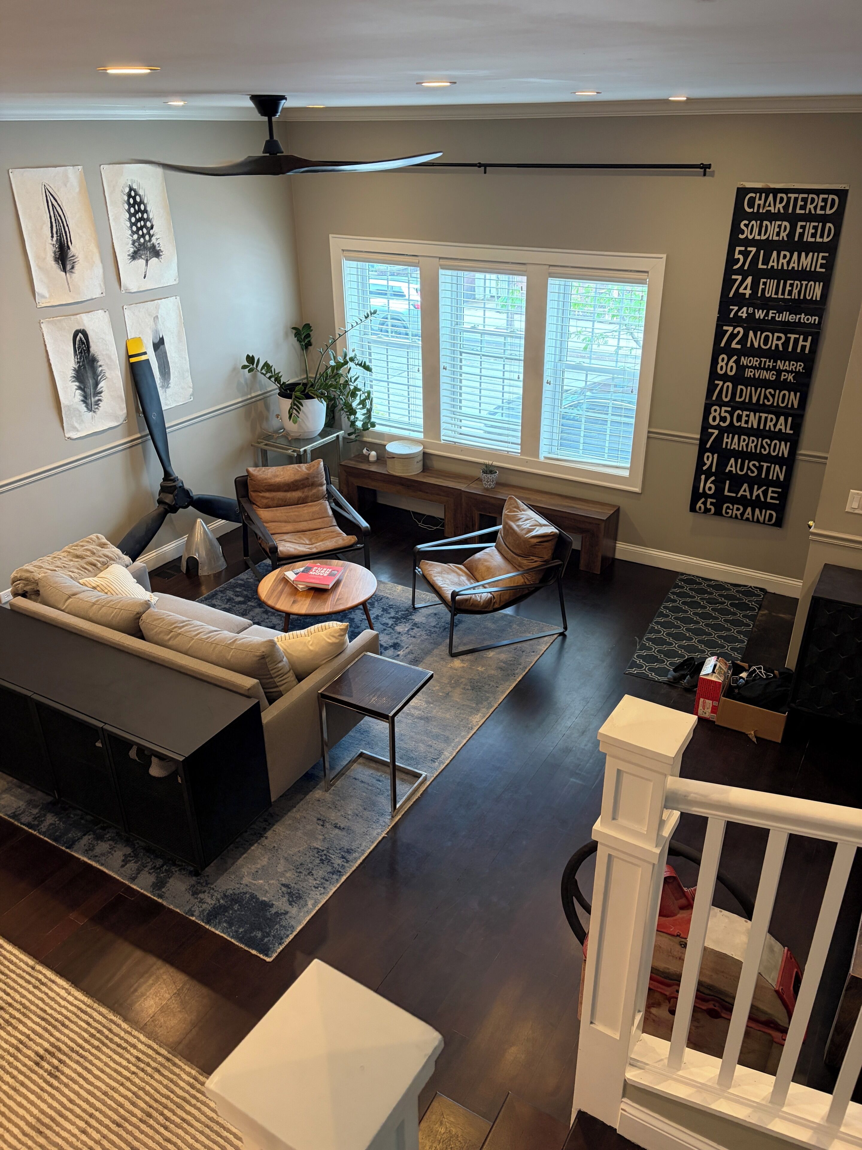 A modern living room with a sectional sofa, a coffee table, and a dining area with a bench. There are large windows providing natural light, and a wall-mounted sign with address information.