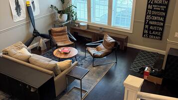 A modern living room with a sectional sofa, a coffee table, and a dining area with a bench. There are large windows providing natural light, and a wall-mounted sign with address information.