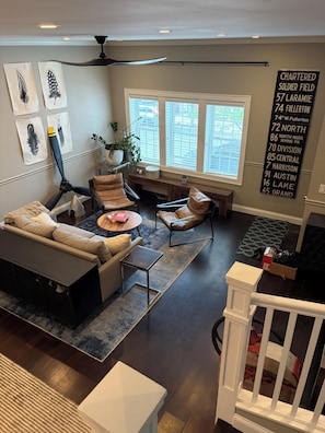 A modern living room with a sectional sofa, a coffee table, and a dining area with a bench. There are large windows providing natural light, and a wall-mounted sign with address information.