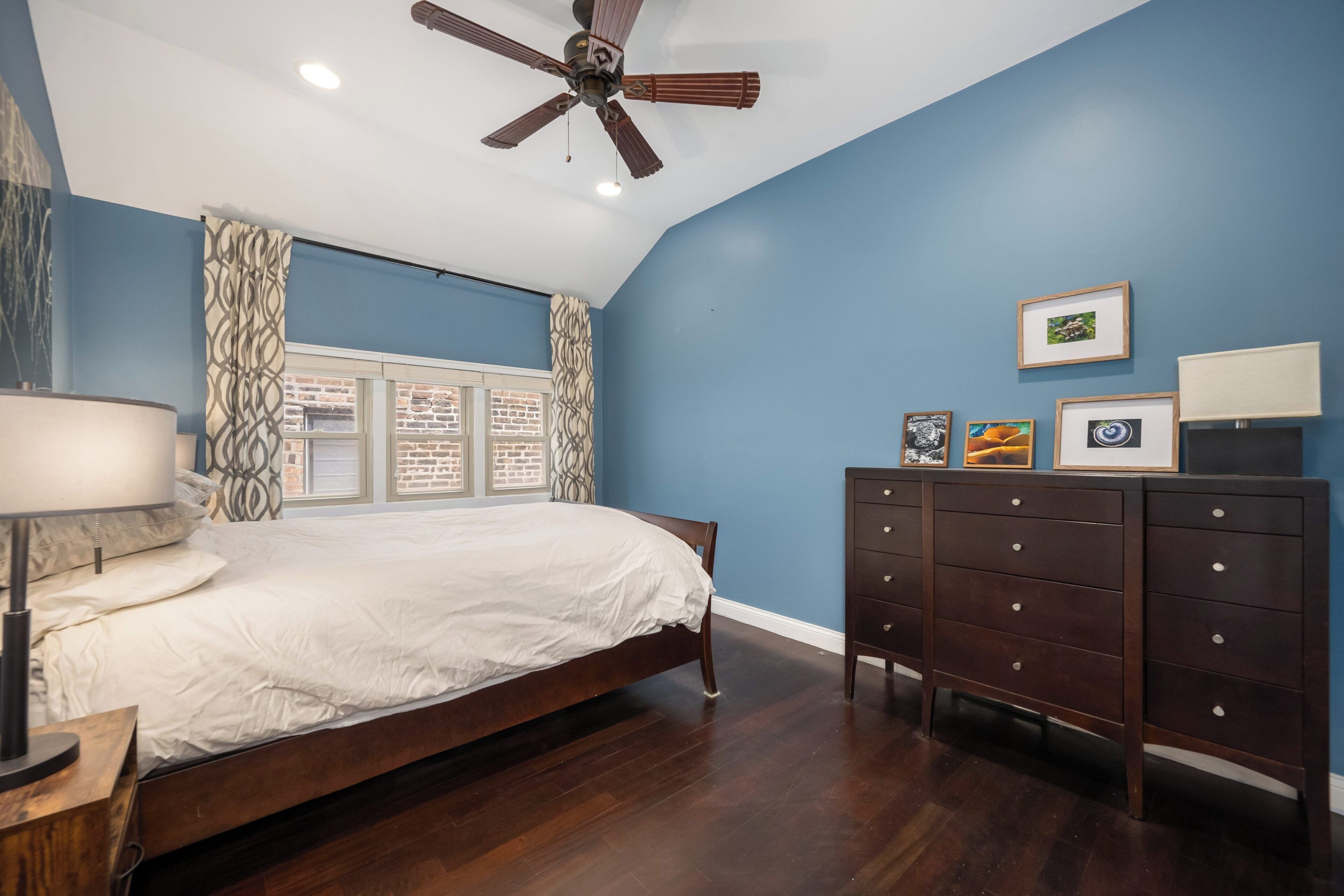 A bedroom with a bed, a dresser, a ceiling fan, and a window with curtains.