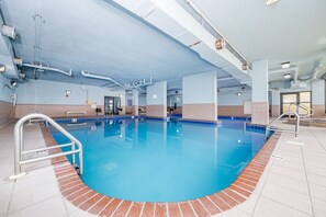 Ein hallenbad mit klarem, blauem Wasser, umgeben von Fliesenböden und -wänden, mit sichtbaren Geländern am Beckenrand und einer Decke mit freiliegenden Rohren und Leuchten.