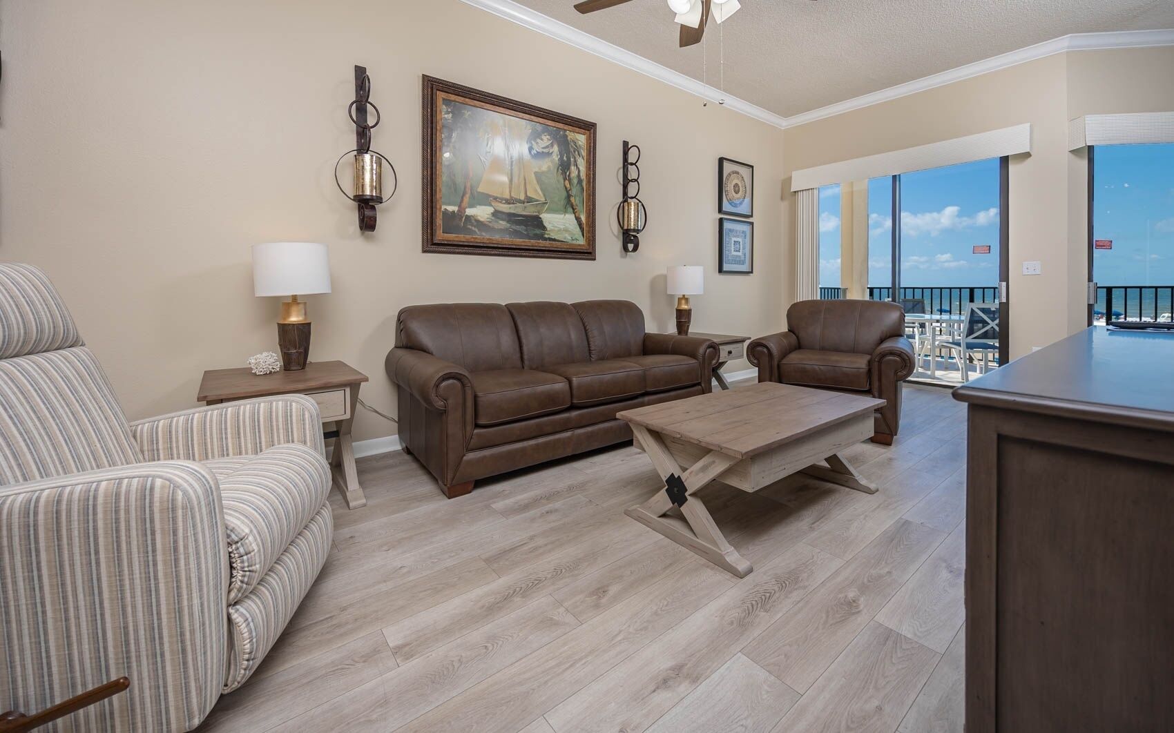 A living room with a brown leather sofa, striped armchair, and a wooden coffee table. There are wall-mounted lamps, a framed painting, and a view of the outside.