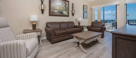 A living room with a brown leather sofa, striped armchair, and a wooden coffee table. There are wall-mounted lamps, a framed painting, and a view of the outside.
