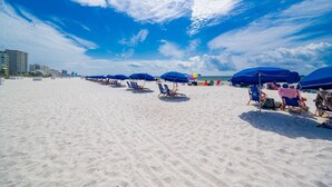 A beach with rows of blue umbrellas and lounge chairs, a clear sky, and buildings in the background. - Phoenix V 111 1 Bedroom Condo (Orange Beach)