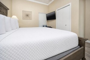 A neatly made bed with a quilted white cover, a bedside lamp, a wall-mounted TV, and a door to the right. - Phoenix V 111 1 Bedroom Condo (Orange Beach)