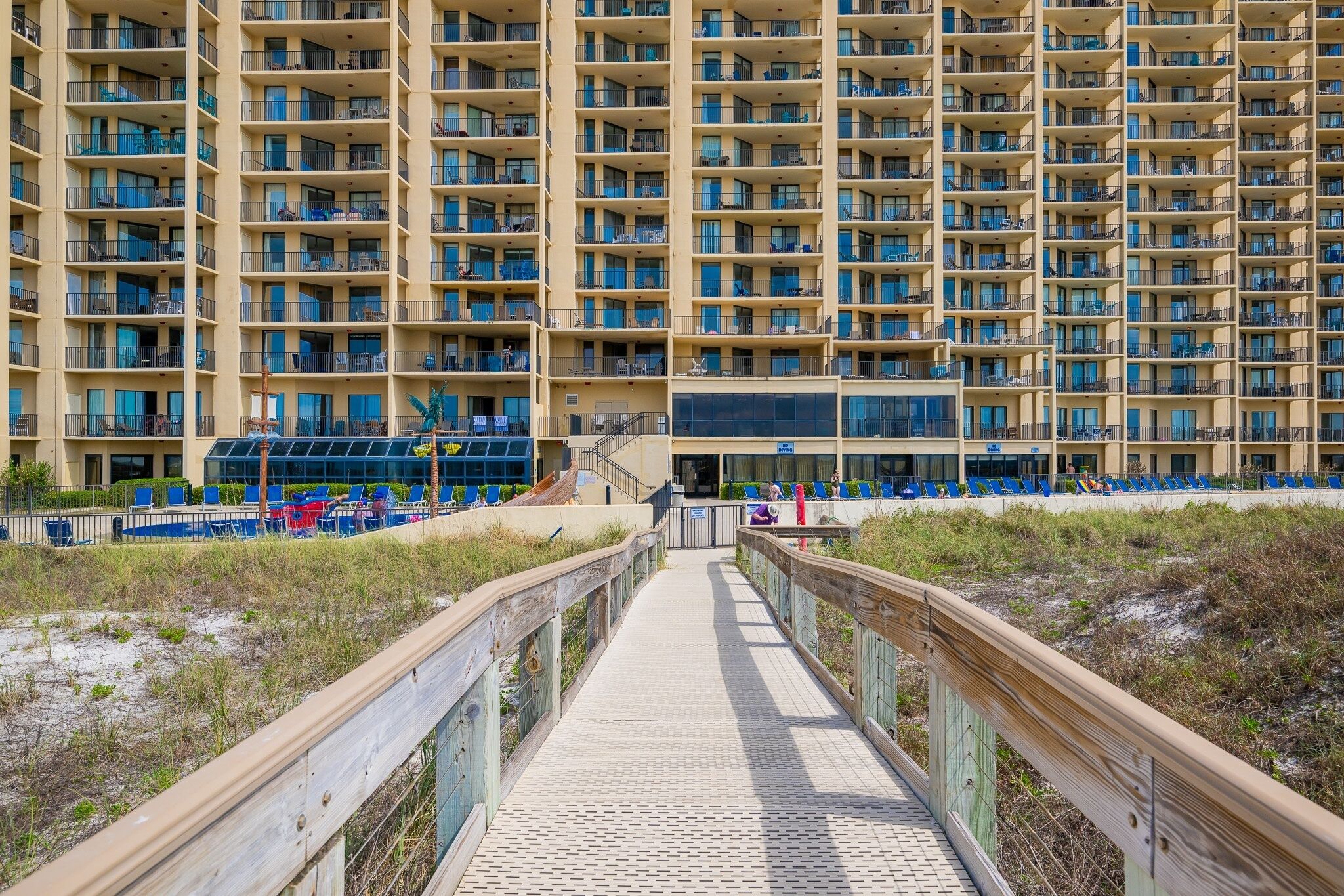 Ein Hochhaus-Hotel mit mehreren Balkonen, einer Strandpromenade und einem Poolbereich mit blauen Liegestühlen.