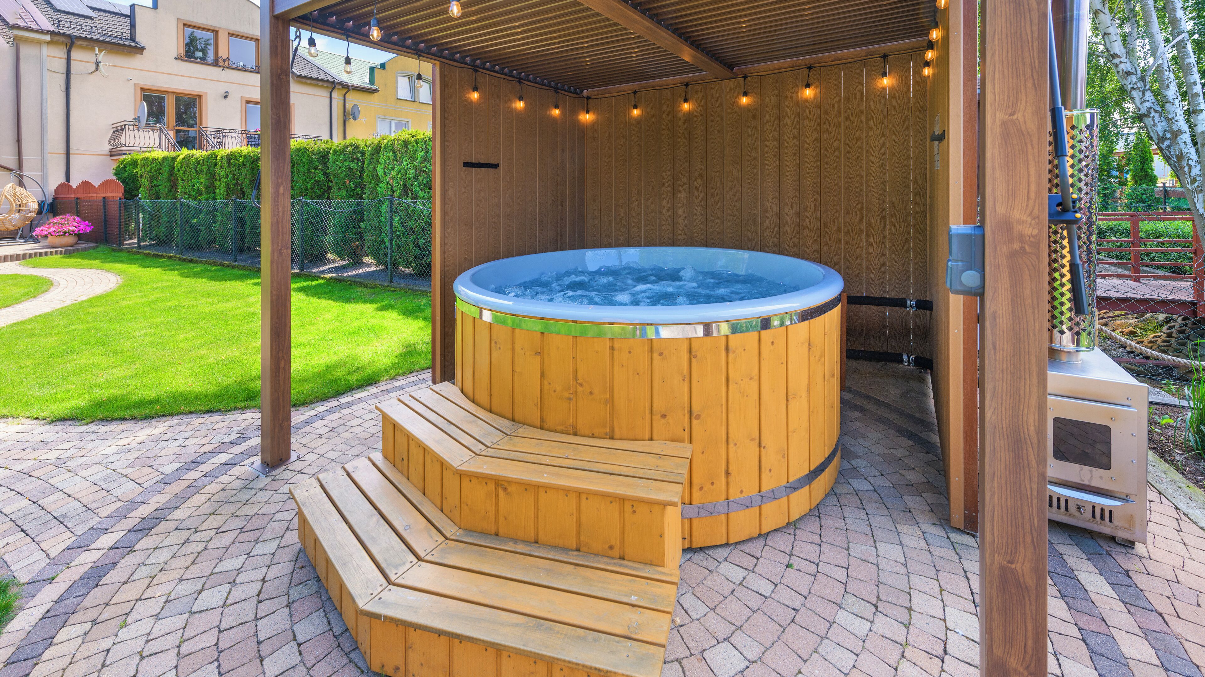 A wooden hot tub with steps, surrounded by a brick patio and a wooden pergola with string lights.