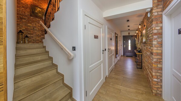 Una escalera de madera conduce a un rellano con una puerta blanca, un muro de ladrillo y un pasillo con piso de madera.