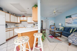 Une cuisine moderne avec des meubles blancs et un coin bar, un salon avec un canapé bleu et un tableau, ainsi qu’un ventilateur de plafond.