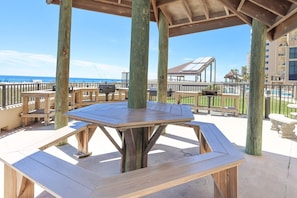 Una terraza de madera con mesas de picnic y una parrilla, con vistas a la playa y una zona de piscina.