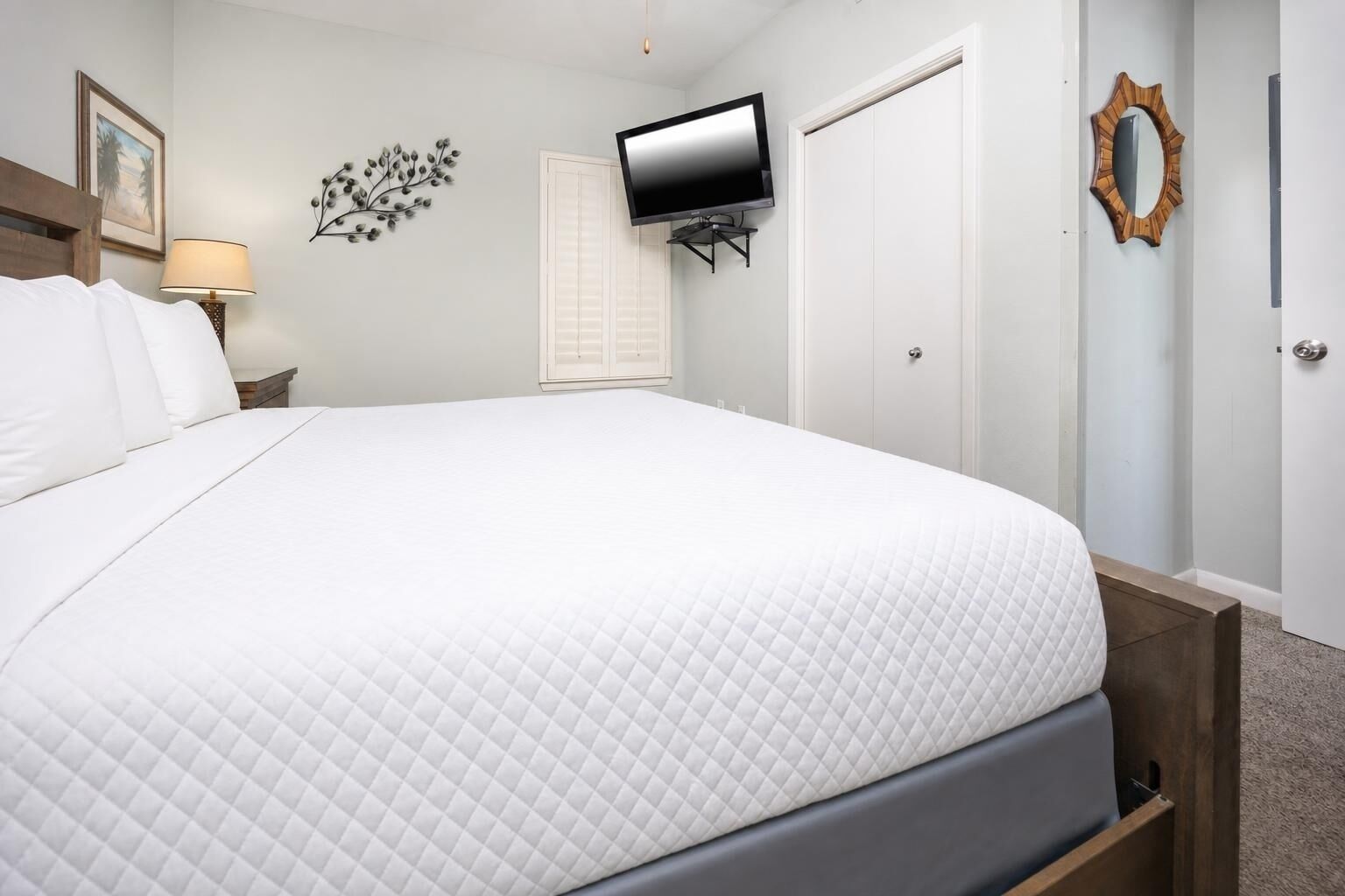 A neatly made bed with a quilted white duvet, a flat-screen TV mounted on the wall, a wooden-framed mirror, and a nightstand with a lamp.