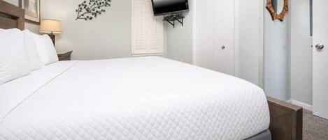 A neatly made bed with a quilted white duvet, a flat-screen TV mounted on the wall, a wooden-framed mirror, and a nightstand with a lamp.