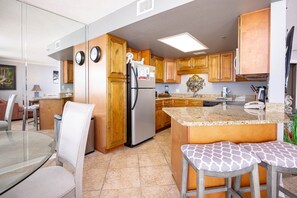 A kitchen with wooden cabinets, a refrigerator, a granite countertop, and a dining area with a glass table and chairs. - Phoenix V 914 1 Bedroom Condo (Orange Beach)