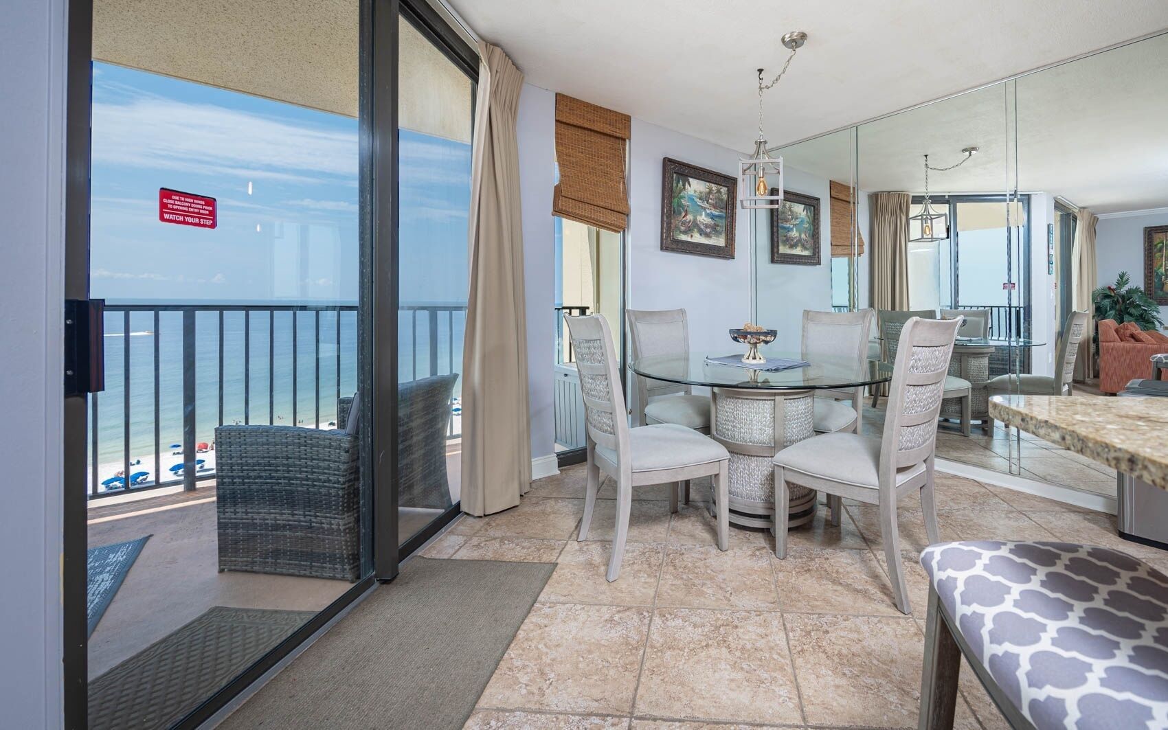 En balkon med en glasdør, der giver udsigt til stranden, et rundt glastabel med stole og en sofa med et mønstret pude.