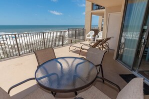 Ein Balkon mit Glastisch und Stühlen, der Blick auf einen Strand mit Zaun und Liegestühlen bietet.
