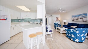 Eine moderne Küche mit weißen Schränken, einer zentralen Kochinsel mit Barhocker und einem Essbereich mit blauem Sofa und Hocker mit Sternenmuster.