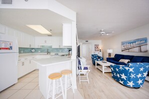 Eine moderne Küche mit weißen Schränken, einer zentralen Kochinsel mit Barhocker und einem Essbereich mit blauem Sofa und Hocker mit Sternenmuster.
