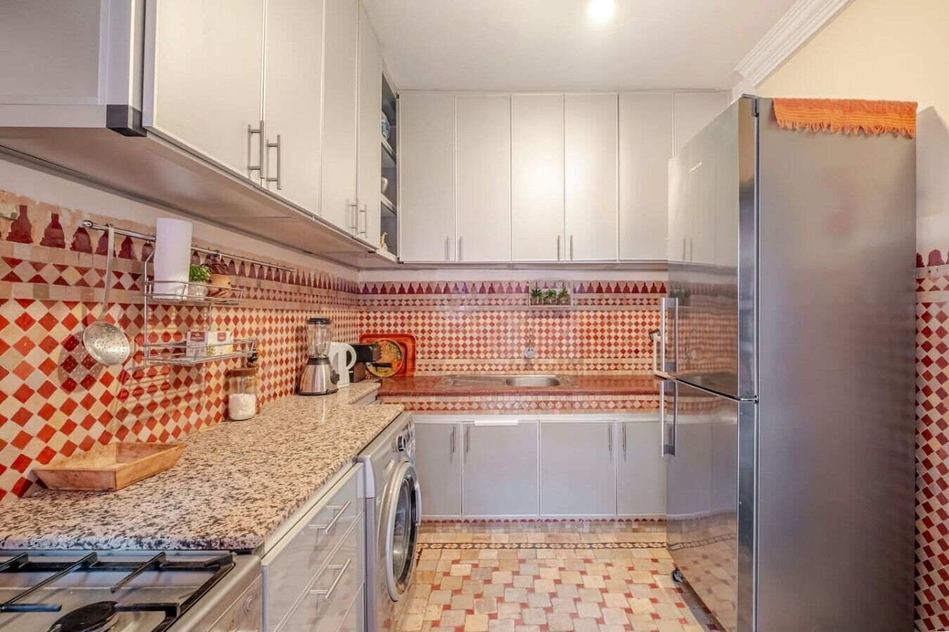 Una cucina compatta con mobili bianchi, un piano di lavoro in granito, un frigorifero in acciaio inossidabile e una parete di fondo rivestita di piastrelle decorate.