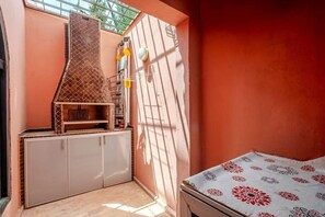 A room with a kitchenette, a bed with a patterned bedspread, and a window with a metal grill. - 3ch, piscine proche Médina, résidence sécurisée (Marrakesh)