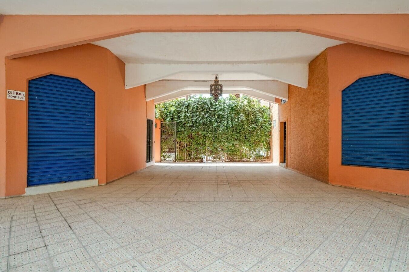 A corridor with closed blue shutters, a tiled floor, and a view of a green tree-lined area through an open gate.