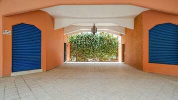 A corridor with closed blue shutters, a tiled floor, and a view of a green tree-lined area through an open gate.