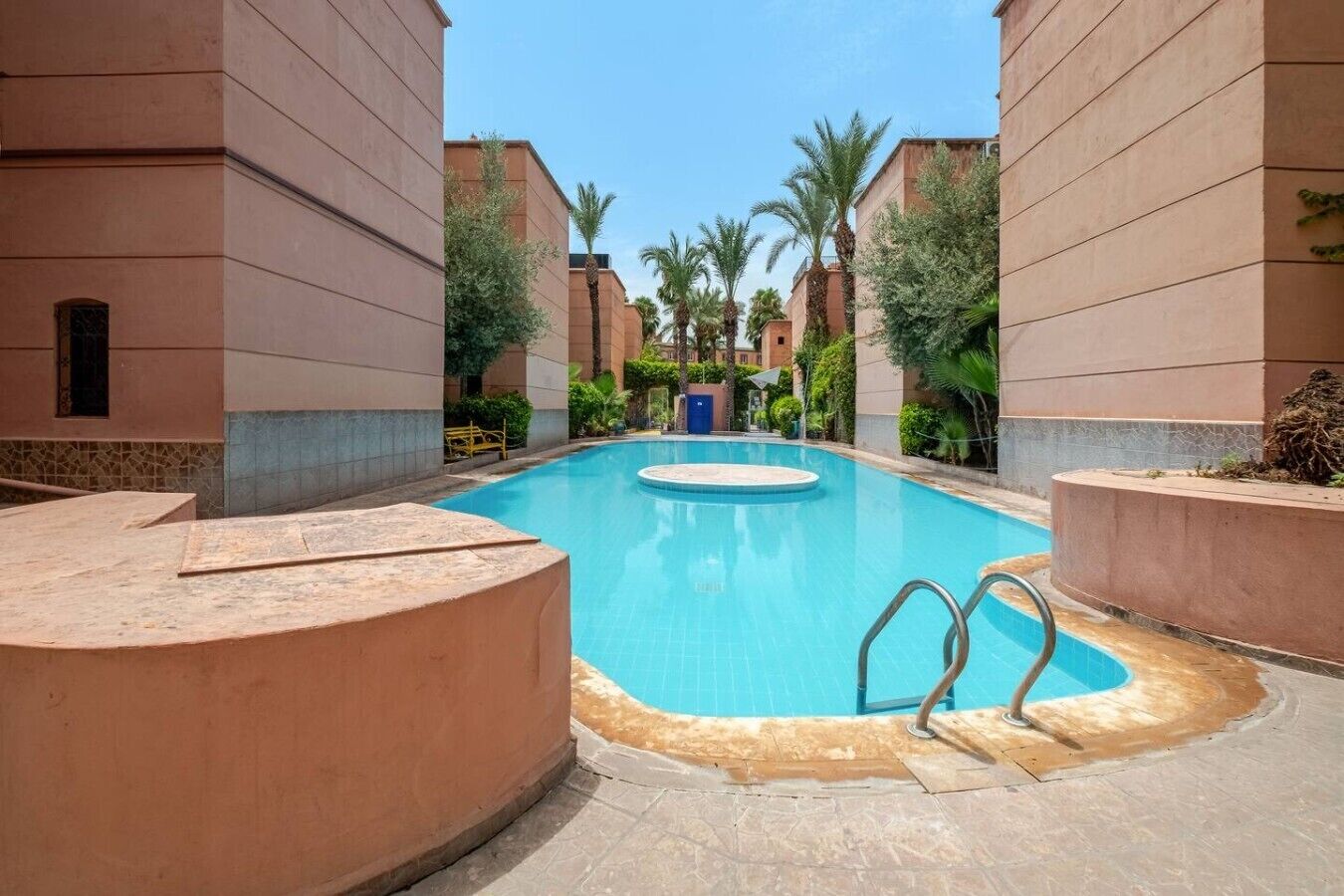 Una piscina con un’area centrale circolare, circondata da palme e edifici con uno stile architettonico uniforme.
