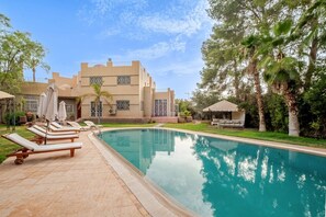 A pool area with lounge chairs and umbrellas, a building with decorative windows, and palm trees. - Villa Piscine / Hammam / 10 min de Centre (Marrakesh)
