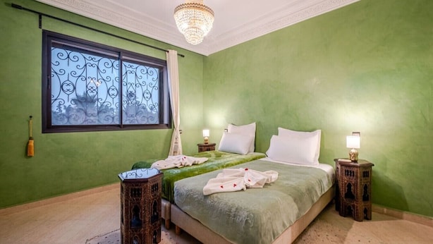 Une chambre avec un mur vert, un grand lit avec une literie blanche, deux tables de chevet avec des lampes, un lustre décoratif et une fenêtre ornée de ferronnerie.