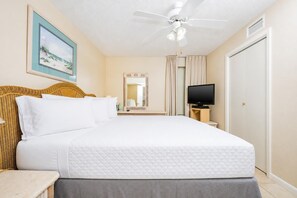 Quarto de hotel com cama, televisão, ventoinha no teto, quadro pendurado na parede e uma porta.