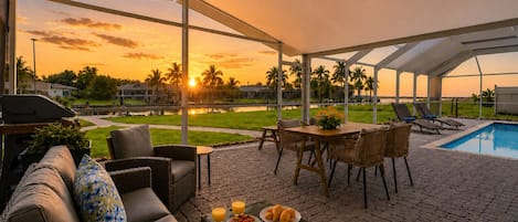 An outdoor patio area with a pool, lounge chairs, a dining table with chairs, and a view of the sunset over a body of water.