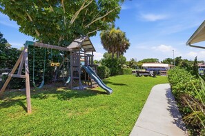 A playground with a slide and swings, surrounded by grass and trees. - Gar4440 2 Bedroom Home (Port Charlotte)
