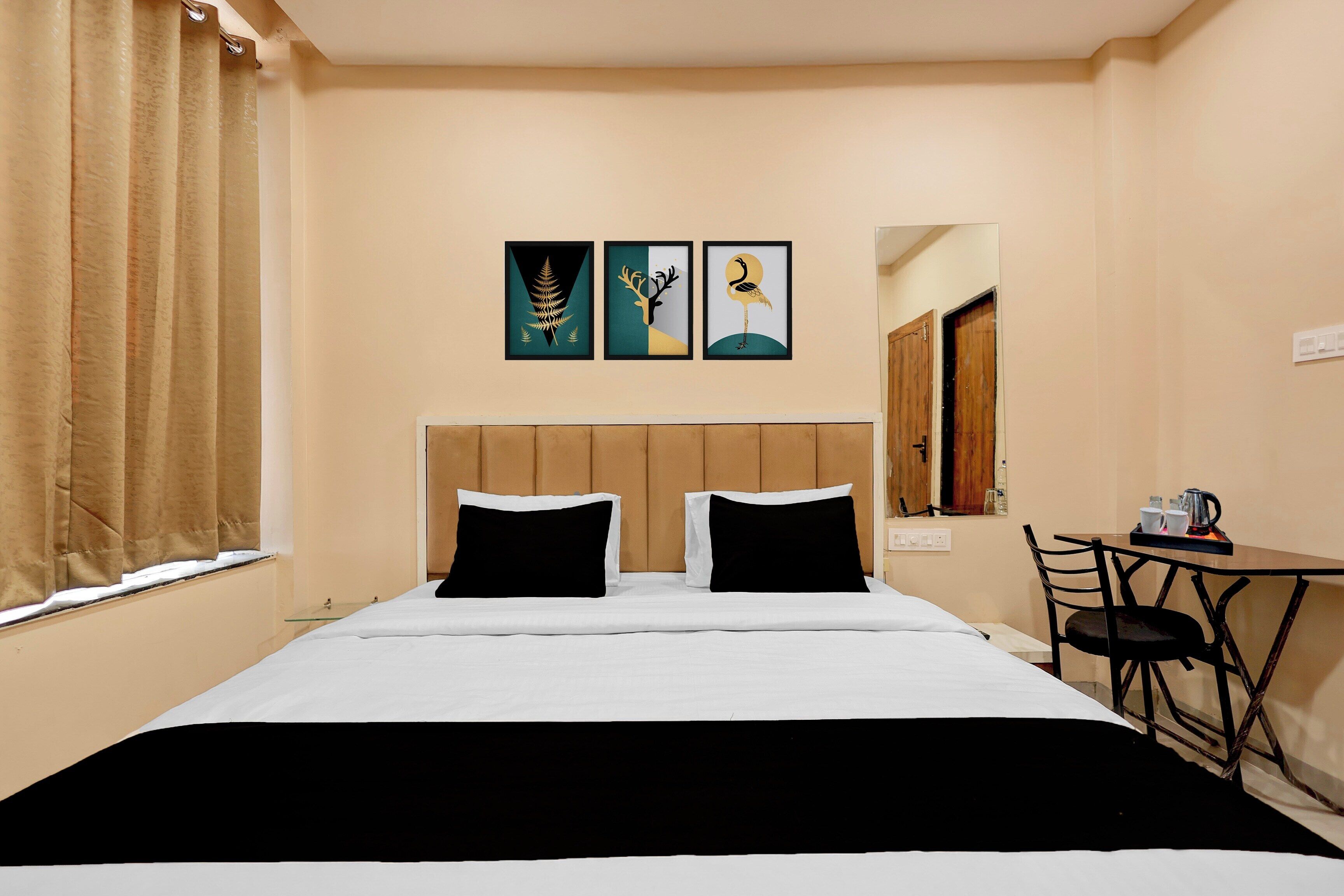 A hotel room with a bed, two black pillows, a wooden headboard, a small table with a chair, and three framed artworks on the wall.