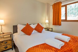 Quarto de hotel com cama, duas almofadas, mesa de cabeceira, abajur, janela com cortinas e vista para árvores.
