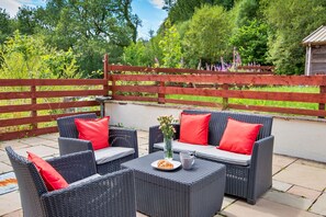 Een terras met rieten meubels en rode kussens, een bord met eten, en een vaas met bloemen.