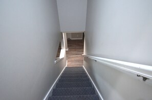 Uma escada estreita com corrimão, que dá acesso a um quarto com soalho de madeira.